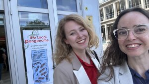 Bia Walter Costa and Rose Brouns in Alicante. In the background a door with the poster of the event "The local Pangenomeal Pangenome"